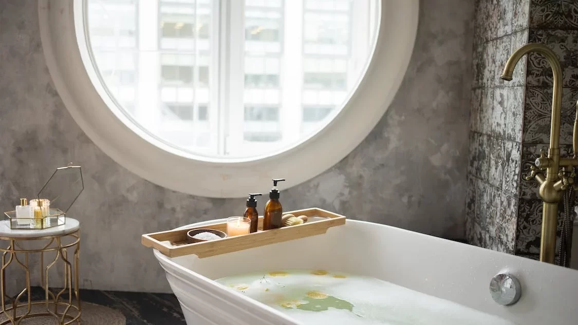 Freestanding bathtub filled with water beside a round window, featuring bath tray, candles, and soft natural light